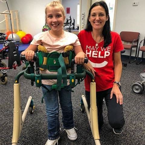 Physical therapist Lisa Davison with a young female patient using a mobility aid. Lisa is wearing a May We Help shirt.